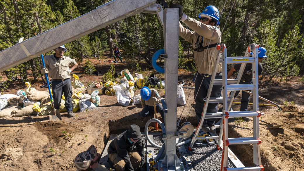 A CCC Corpsmember stands on a ladder, helping project sponsors install a remote weather station.