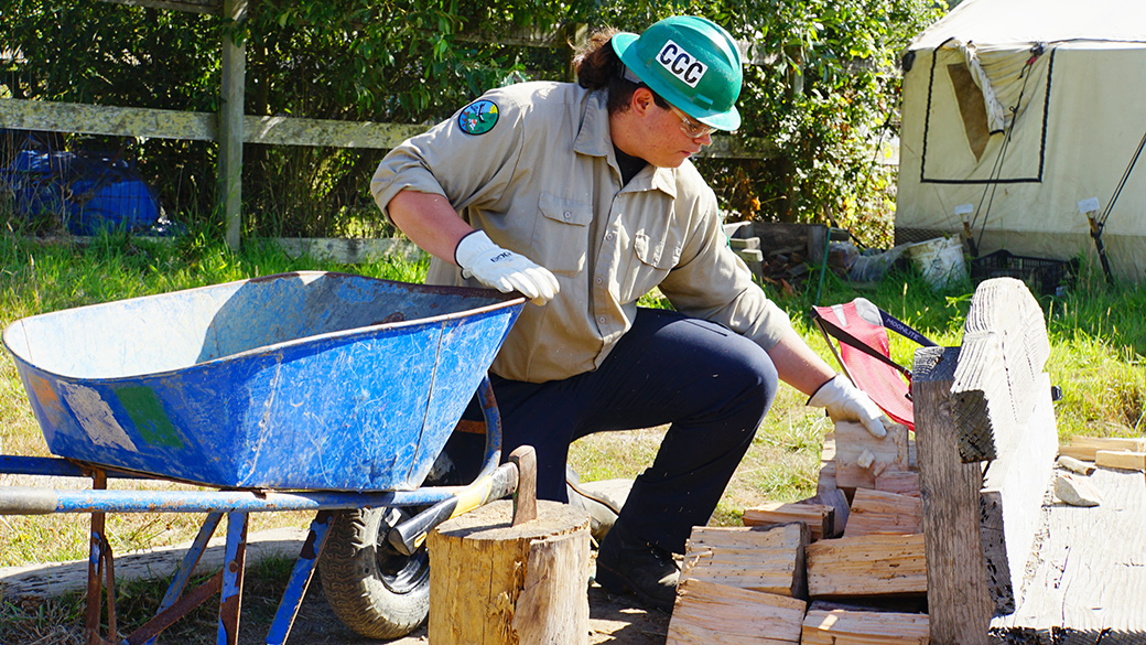 male in green helmet unloading wood from wheelbarrow near camp fire