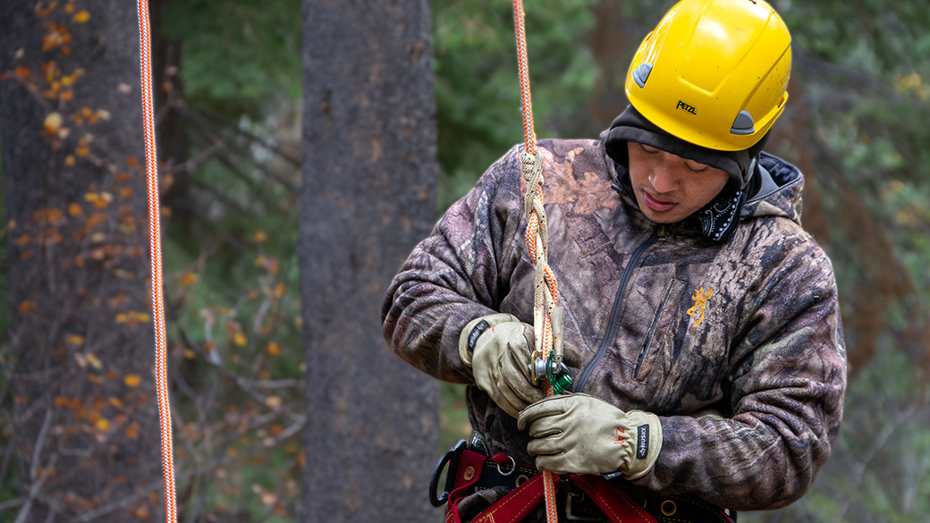male in safety harness and helmet manipulates a caribbeaner in a forest