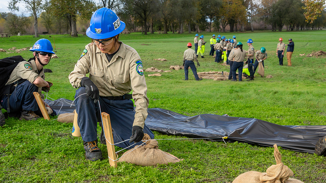 female corpsmember tying twine around stake for sandbag with visqueen in the background