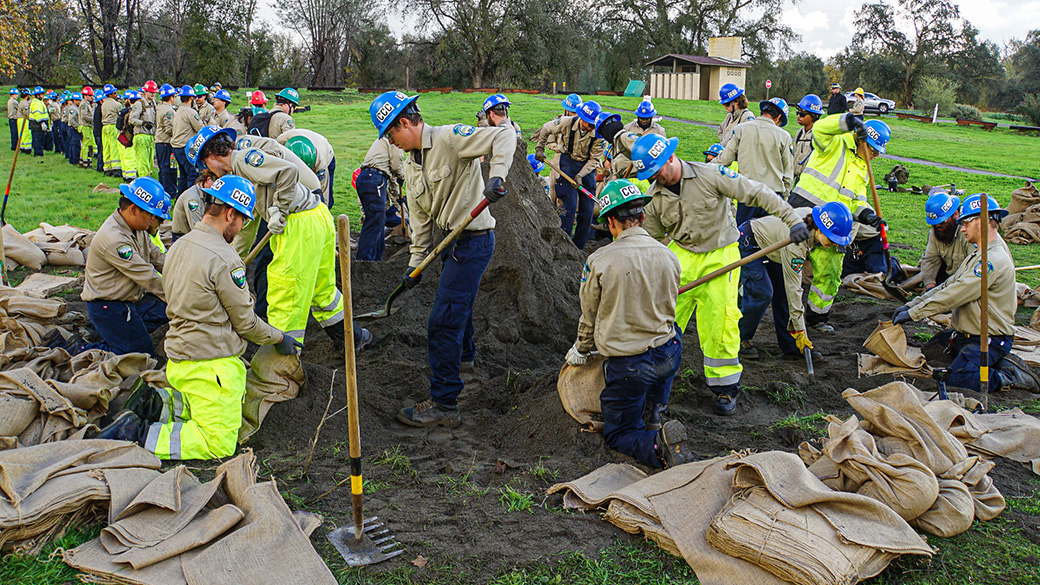 corpsmembers stand and kneel around a large pile of sand to fill sandbags in a park