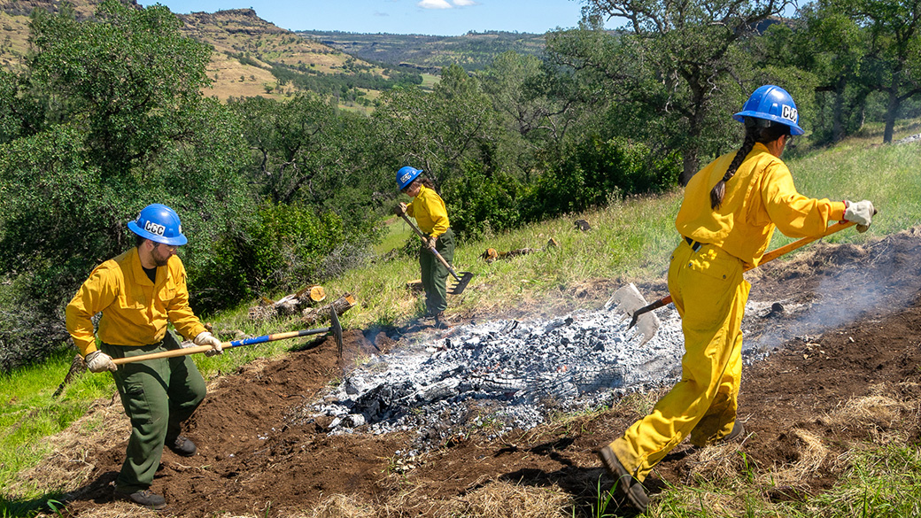 three corpsmembers use hoses and tools around burned wood and charcoal