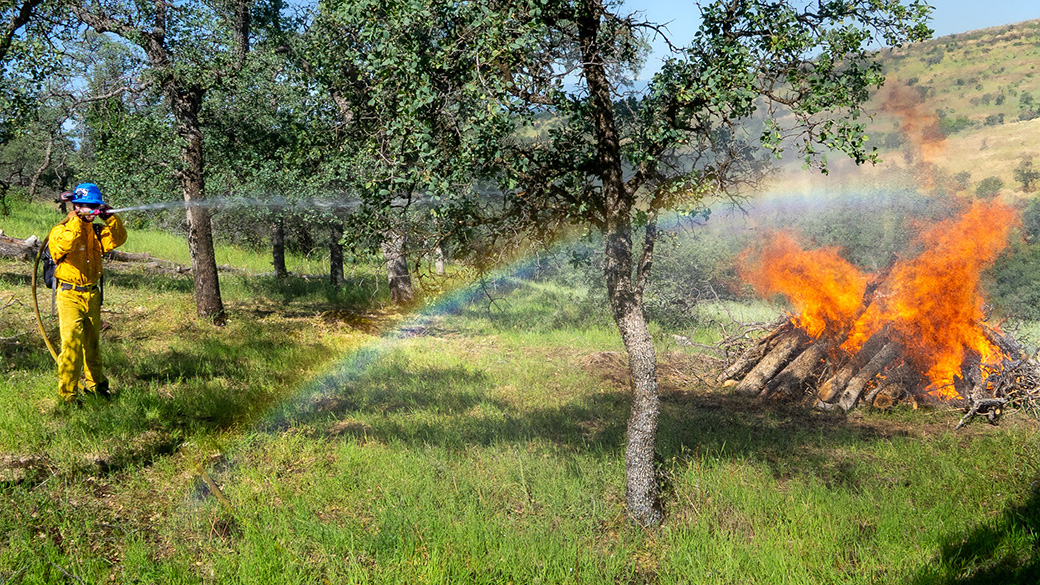 A Corpsmember in yellow nomex uses a fire hose to control a burning wood pile which forms a rainbow near the flames.