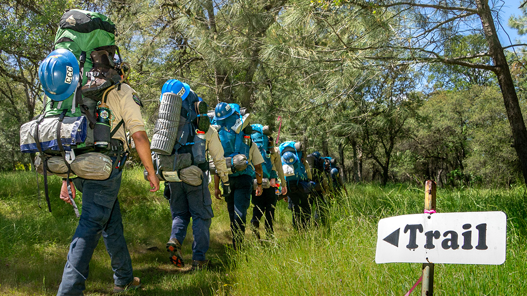 corpsmembers with backpacks on hike past green grass and a sign with an arrow that says 'trail'